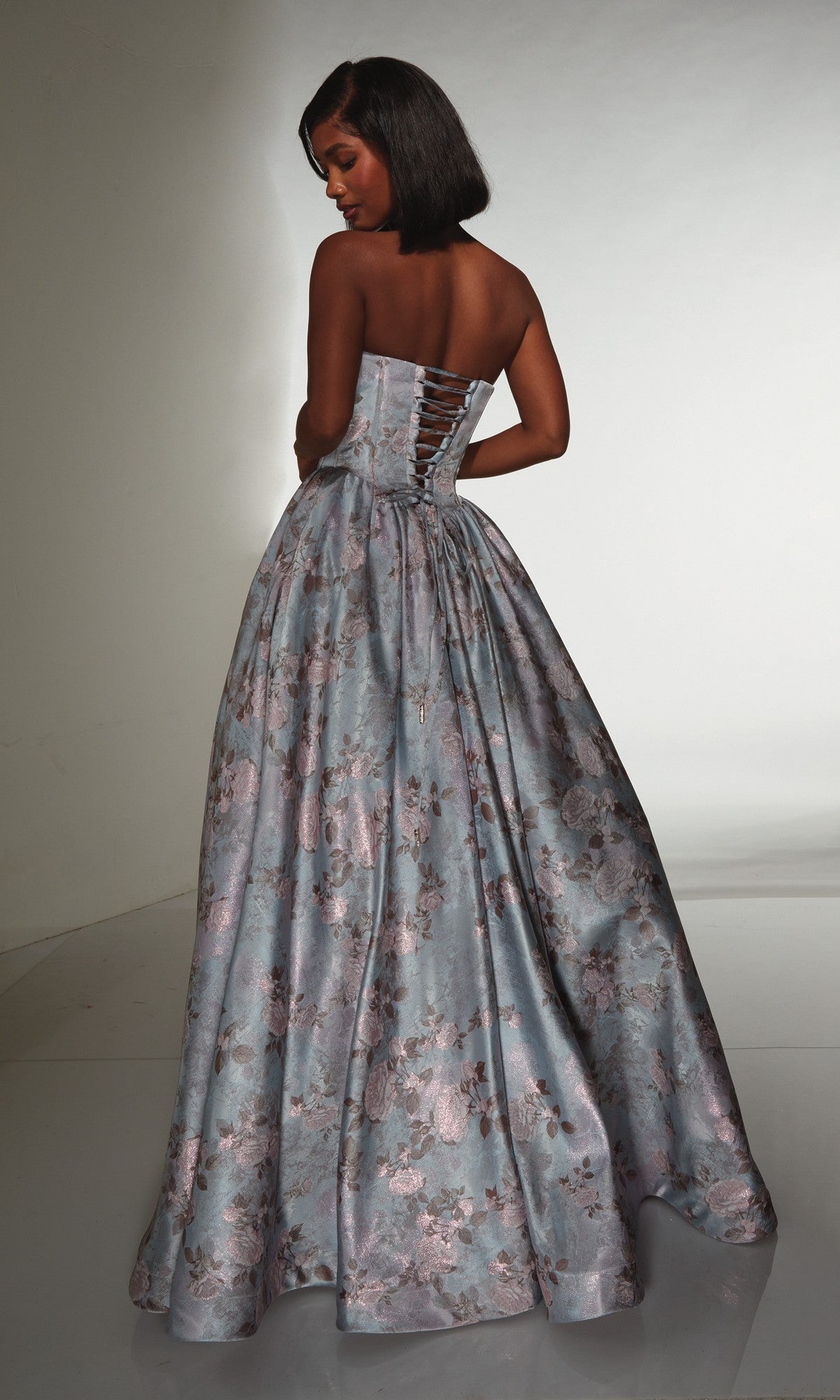 A woman stands with her back to the camera in the Alyce 62181 long prom dress, a strapless ball gown with a lace-up corset bodice and floor-length floral brocade design, set against a plain background.